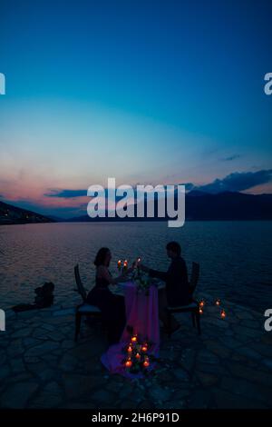 Mann und Frau sitzen an einem Tisch und klirken nachts auf dem Pier vor dem Hintergrund des Meeres und der Berge, indem sie Kerzen verbrennen Stockfoto