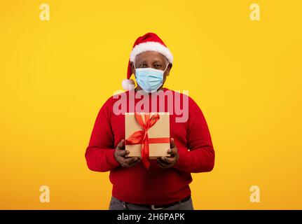 Weihnachten und Neujahr während der Corinavirus-Sperre. Älterer schwarzer Mann in Schutzmaske und Weihnachtsmann Hut mit Geschenkbox Stockfoto