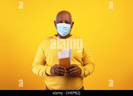 Älterer schwarzer Mann in medizinischer Maske mit Flugtickets und internationalem Pass auf orangenen Studiohintergrund Stockfoto