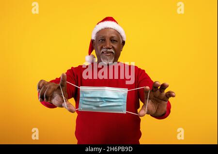 Glücklicher älterer schwarzer Mann im Weihnachtsmann-Hut, der Gesichtsmaske in ausgestreckten Händen hält und Weihnachten ohne Coronavirus feiert Stockfoto