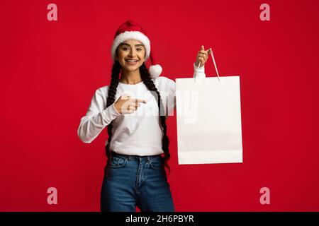 Glückliche Arabische Frau In Santa Hat Zeigt Auf Blank Paper Shopping Bag Stockfoto