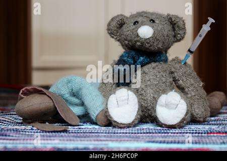 Zwei Kuscheltiere, ein liegender Hase und ein sitzender Teddy mit Spritze im Arm auf einem bunten Teppich. Stockfoto