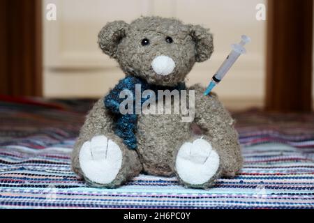 Kuscheltier Teddy mit Spritze im Arm, sitzend auf einem bunten Teppich. Stockfoto