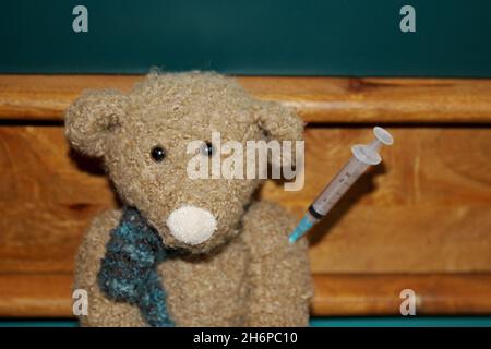 Kuscheltier Teddy mit Spritze im Arm, sitzend vor einem braun grünen Hintergrund. Stockfoto