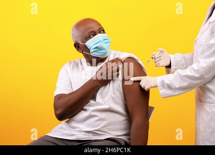 Arzt, der einen Impfstoff gegen das Coronavirus bei einem älteren schwarzen männlichen Patienten auf orangefarbenem Studiohintergrund abschoss Stockfoto