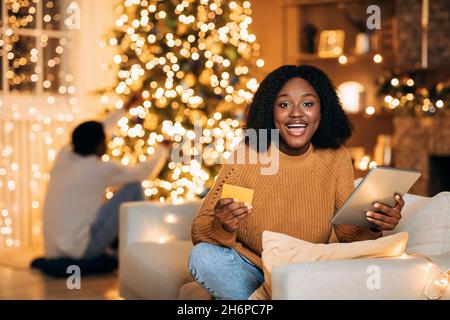 Schwarze Frau mit Tablet und Kreditkarte, Einkaufen für Weihnachtsgeschenke online, während ihr Freund Dekoration Tannenbaum zu Hause Stockfoto