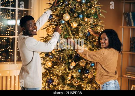 Glückliches junges schwarzes Paar, das zu Hause einen schönen Weihnachtsbaum schmückt Stockfoto