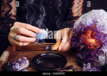 Glamouröse Frau im schwarzen Kleid reinigt Kristalle Edelsteine durch verschmierenden Palo Santo Holzstab. Entfernen Sie negative Energie. Stockfoto