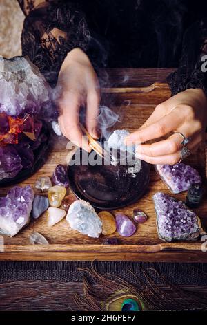 Glamouröse Frau im schwarzen Kleid reinigt Kristalle Edelsteine durch verschmierenden Palo Santo Holzstab. Entfernen Sie negative Energie. Stockfoto