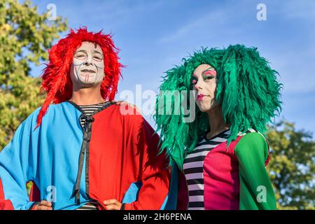 Positive männliche und weibliche Harlekine mit gemalten Gesichtern in bunten Kostümen und Perücken, die an einem sonnigen Tag lächeln. Stockfoto