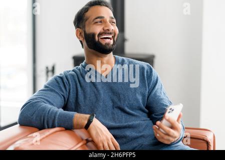 Urkomischer Indianer aus den 30er Jahren, der Smartphone benutzt und zu Hause auf der Couch sitzt, hält ein lateinamerikanischer Mann das Handy und lacht fröhlich, schaut sich Komödien an, schaut lustige Videos an und macht mit Freunden online Scherz Stockfoto