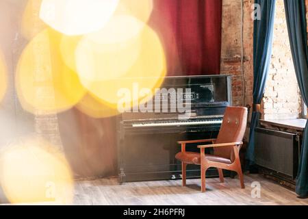 Altes Vintage-Klavier und Sessel stehen in der Halle vor dem Hintergrund eines roten Vorhangs und einer Backsteinmauer in der Nähe des großen Fensters, mit Bokeh und Highlig Stockfoto