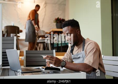 Junger Mann mit Schürze sitzt am Tisch vor dem Laptop in einem modernen Café und nimmt Online-Bestellungen von Kunden zur Lieferung entgegen Stockfoto