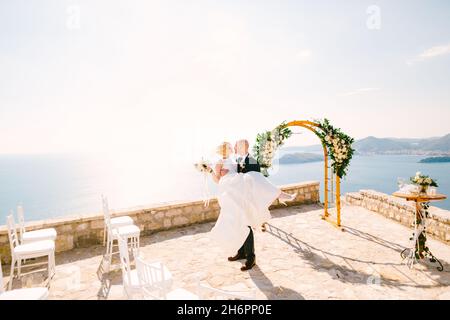 Bräutigam hält die Braut in einem weißen Kleid mit einem Blumenstrauß in den Armen, während sie auf der Aussichtsplattform mit Blick auf den Sveti Stefan steht Stockfoto