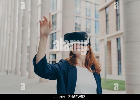 Fröhliche Rotschopf-Frau, formell gekleidet, steht im Freien in einer Virtual-Reality-Brille Stockfoto