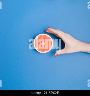 Weibliche Hand mit roten Nägeln, die in Richtung einer hellen Grapefruitscheibe reichen. Gesunde saftige Fruchtzusammensetzung. Stockfoto