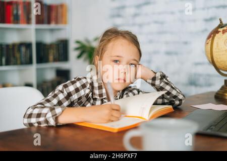 Vorderansicht der gelangweilten müde niedlichen Grundschule Kind Schulmädchen tun Schreiben Hausaufgaben halten Stift sitzen am Tisch mit Laptop und Papier-Notebook zu Hause Stockfoto