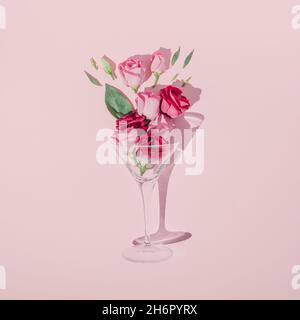 Romantische Komposition zum Valentinstag mit Cocktailglas, grünen Blättern und rosa Rosen auf pastellfarbenem Hintergrund. Minimale flache Verlegung. Stockfoto