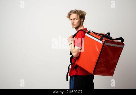 Angestellter Mann in einem roten T-Shirt Uniform Arbeitskleidung arbeiten als Händler Kurier und halten roten thermischen Lebensmittelbeutel Rucksack isoliert auf weiß Stockfoto