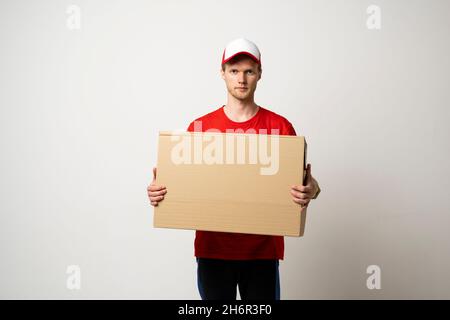 Männlicher Angestellter in roter Uniform, der als Kurierhändler arbeitet, hält den Karton isoliert auf weißem Hintergrund, Studioportrait. Servicekonzept. Mock Stockfoto