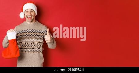 Konzept der Winterferien, Neujahr und Feier. Fröhlich schöner Mann in weihnachtsmann Hut und Pullover, zeigt weihnachtsstrumpf mit Süßigkeiten und Stockfoto