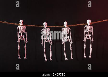 Vier Skelette, die am Hals an einem Seil mit Silhouetten auf dunklem Hintergrund hängen. Halloween-Dekoration Stockfoto