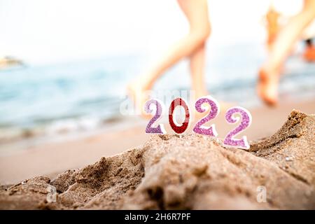 Zahlen 2022 auf Sand vor dem Hintergrund des Meeresstrandes im Resort in südlichen Ländern mit Füßen von Urlaubern, die bei Sonnenuntergang und Celebrati auf dem Wasser spazieren Stockfoto