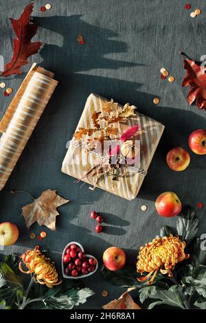 Orangefarbene Chrysanthemenblüten, trockenes Hasenschwanzgras, Kürbisse, Cranberry- und rote Eichenblätter. Eingepacktes Geschenk in der Hand. Herbstschmuck, Draufsicht im Dunkeln Stockfoto