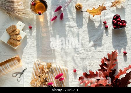 Herbstdekorationen - Preiselbeere, Kekse. Tee und trockene Eichenblätter. Flach liegend, Draufsicht auf unfarbigem Textil. Fallenlassen Hintergrund mit Copy-Space. Stockfoto