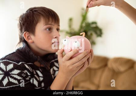 Junge mit einem Sparschwein, der um Geld bettelt. Hand werfen eine Münze in ein Sparschwein. Teenager verlangen Geld, um Geld zu sparen. Persönliche Ausgaben der Schule Stockfoto