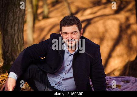 Junger Mann im Mantel Porträt im Herbstwald mit Bäumen Schatten auf dem Hintergrund Stockfoto