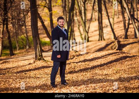 Junger Mann im Mantel Porträt im Herbstwald mit Bäumen Schatten auf dem Hintergrund Stockfoto