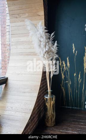 Getrocknete Blumen aus Schilf und hohem Gras in einer hohen Glasvase an einer blauen Wand im Inneren Stockfoto