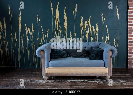 Dunkelgraues gestepptes altes Vintage-Sofa mit Kissen auf einem Holzboden in der Nähe einer dunkelblauen Wand mit bemalten goldenen Ohren Stockfoto