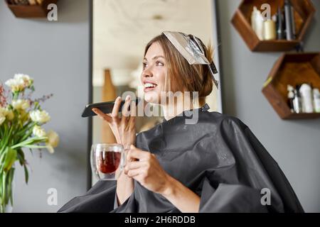 Glückliche blonde kaukasische Frau verwendet Smartphone, während sie im Stuhl sitzt und im Friseursalon wartet Stockfoto