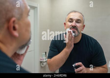 Porträt eines hübschen grauhaarigen Mannes mittleren Alters, der Rasierschaum auftrug, um seinen Bart zu trimmen Stockfoto