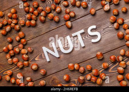 Das Wort Nüsse mit silbernen Buchstaben auf Holzbrett Hintergrund mit verstreuten Haselnüssen umgeben gelegt Stockfoto
