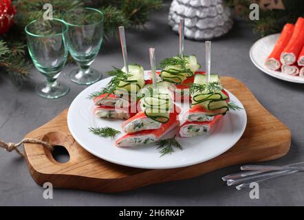 Kanapees mit Krabbenstäbchen, Käse und Gurke auf weißem Teller auf Holzbrett. Festlicher, hausgemachter Snack Stockfoto