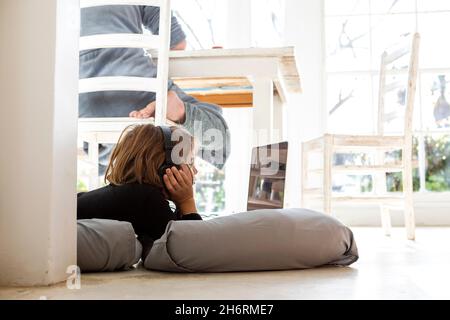 Acht Jahre alter Junge, der auf Kissen liegt, sich an den Händen kinn, einen Laptop-Bildschirm ansieht, Hausaufgaben macht. Stockfoto