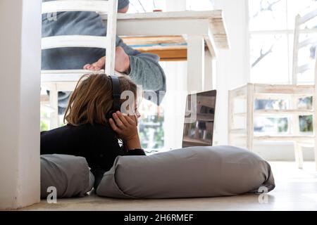 Acht Jahre alter Junge, der auf Kissen liegt, sich an den Händen kinn, einen Laptop-Bildschirm ansieht, Hausaufgaben macht. Stockfoto