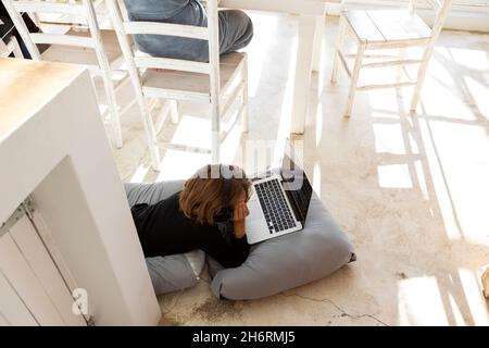 Acht Jahre alter Junge, der auf Kissen liegt, sich an den Händen kinn, einen Laptop-Bildschirm ansieht, Hausaufgaben macht. Stockfoto