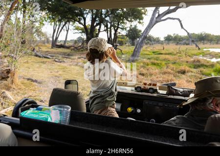 Kleiner Junge mit Ferngläsern, der in einem Safari-Jeep aufsteht und über die Landschaft blickt Stockfoto