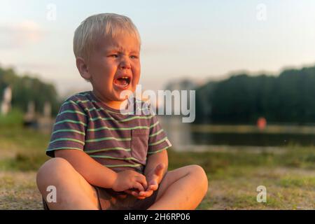 Ein kleiner Junge sitzt auf dem Boden am See im Park und weint heftig. Groll oder Hysterie in einem Kind. Stockfoto