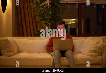 Ein junger Mann mit einem Bart in einem roten Hemd arbeitet mit einem Laptop und sitzt abends im Haus auf der Couch. Stockfoto