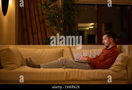 Ein junger Mann mit einem Bart in einem roten Hemd arbeitet mit einem Laptop und sitzt abends im Haus auf der Couch. Stockfoto