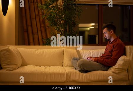 Ein junger Mann mit einem Bart in einem roten Hemd arbeitet mit einem Laptop und sitzt abends im Haus auf der Couch. Stockfoto