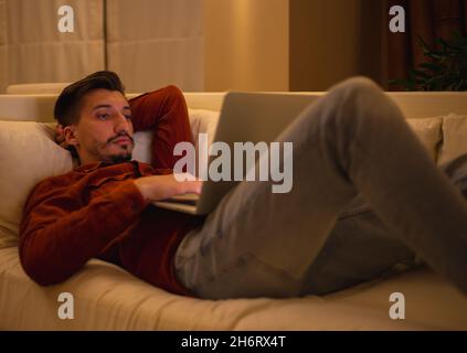Ein junger Mann mit einem Bart in einem roten Hemd arbeitet mit einem Laptop und sitzt abends im Haus auf der Couch. Stockfoto