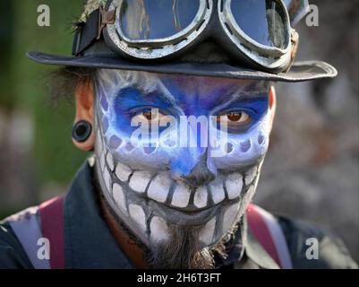 Kostümierter junger mexikanischer Yucatecan-Mann mit farbenfrohem gruseligen Gesichtsbemalung und Ohrstöpseln blickt den Zuschauer am Tag der Toten (Día de los Muertos) an. Stockfoto