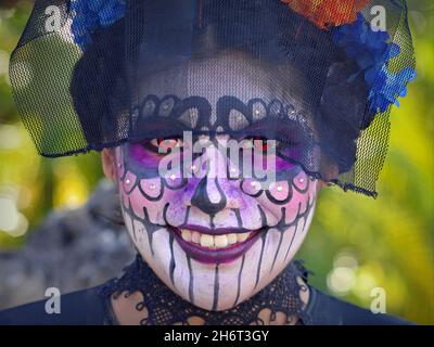 Kostümierte junge, hübsche mexikanische Yucatecan-Frau mit Gesichtsbemalung und orangefarbenen Kontakten lächelt den Betrachter am Tag der Toten (Día de los Muertos) an. Stockfoto