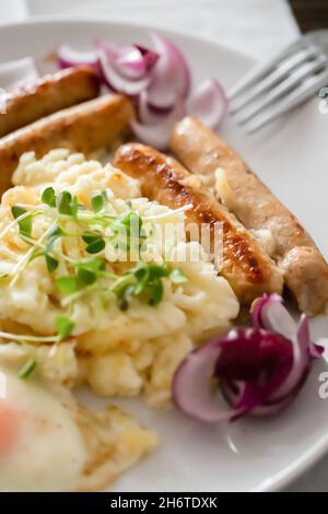 Schweinswürste mit Zwiebeln, Eiern, Kartoffelpüree und Microgreens. Herzhaftes Frühstück. Dunkler Holzhintergrund, weiße Keramikplatte. Draufsicht. Stockfoto